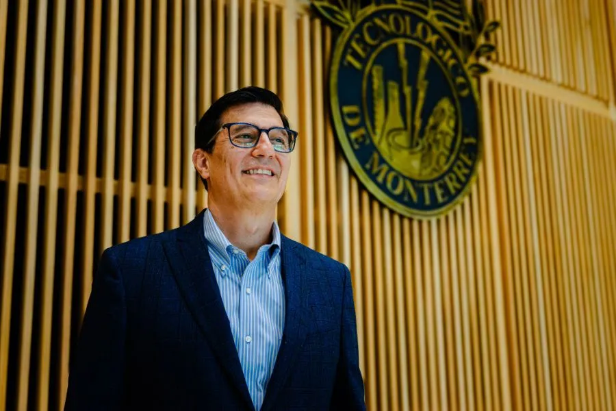 Hugo Garza Medina posa de pie frente al escudo del Tecnol&oacute;gico de Monterrey dentro de un edificio institucional.