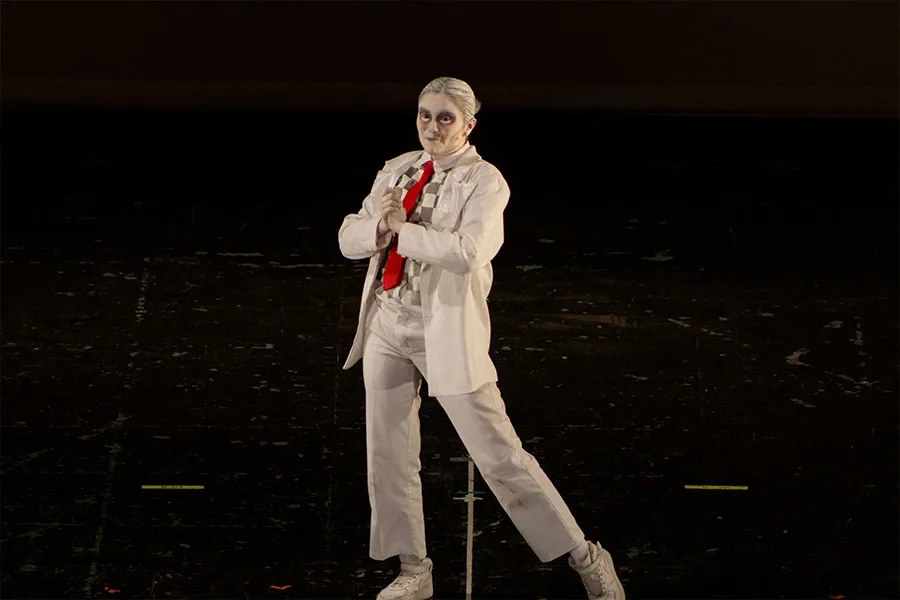 Mujer vestida con pantal&oacute;n, saco blanco, camisa a cuadros blancos y grises y corbata roja en un escenario oscuro, dejando el fondo negro.  