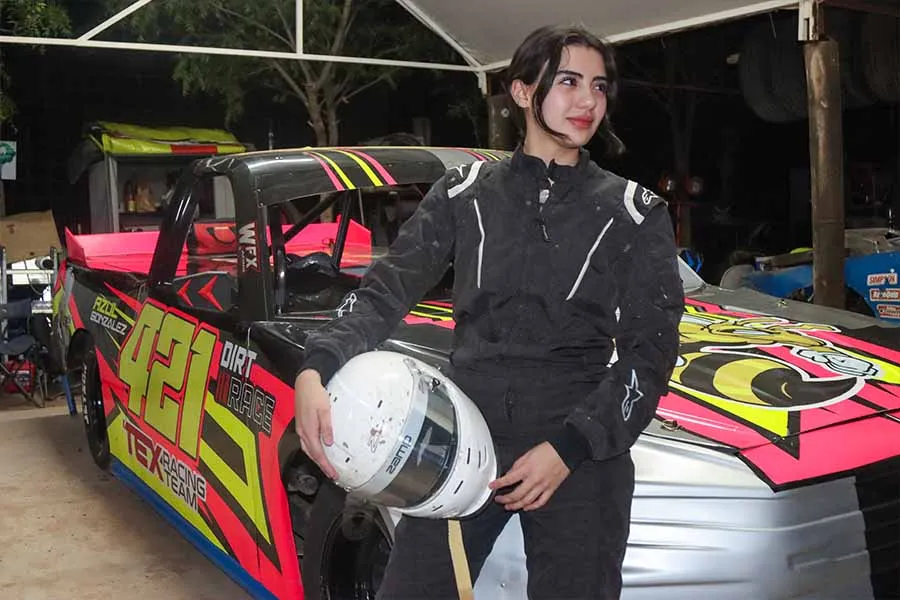 Mujer joven posa en uniforme de piloto automovil&iacute;stico junto a su carro.