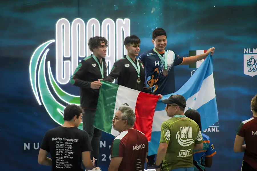 Diego Lizardi, estudiante de PrepaTec y nadador hidalguense, en el podio con la medalla de oro en el CCCAN 2024, sosteniendo la bandera de M&eacute;xico junto a otros medallistas.