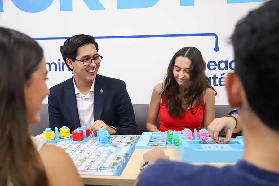 Cuatro estudiantes, dos de espalda y dos de frente, est&aacute;n sentados en una mesa mientras juegan un juego de estrategia. 