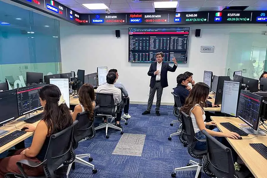 Profesor al frente de un sal&oacute;n, con una pantalla que muestra muchos n&uacute;meros y datos financieros. Frente a el est&aacute;n sentados en dos filas alumnos en estaciones de c&oacute;mputo con pantallas que muestran los mismos n&uacute;meros. 