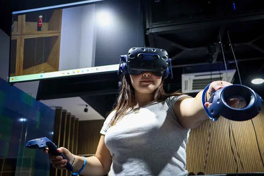 Una chica portando con visor de realidad virtual sostiene en sus manos dos controles. En el fondo cuelga una pantalla del techo que muestra la simulaci&oacute;n que ella experiemnta.