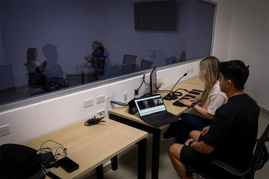 Dos estudiantes est&aacute;n del otro lado de una ventana a trav&eacute;s de la cual se ve un grupo de personas sentadas en una mesa redonda de cristal.