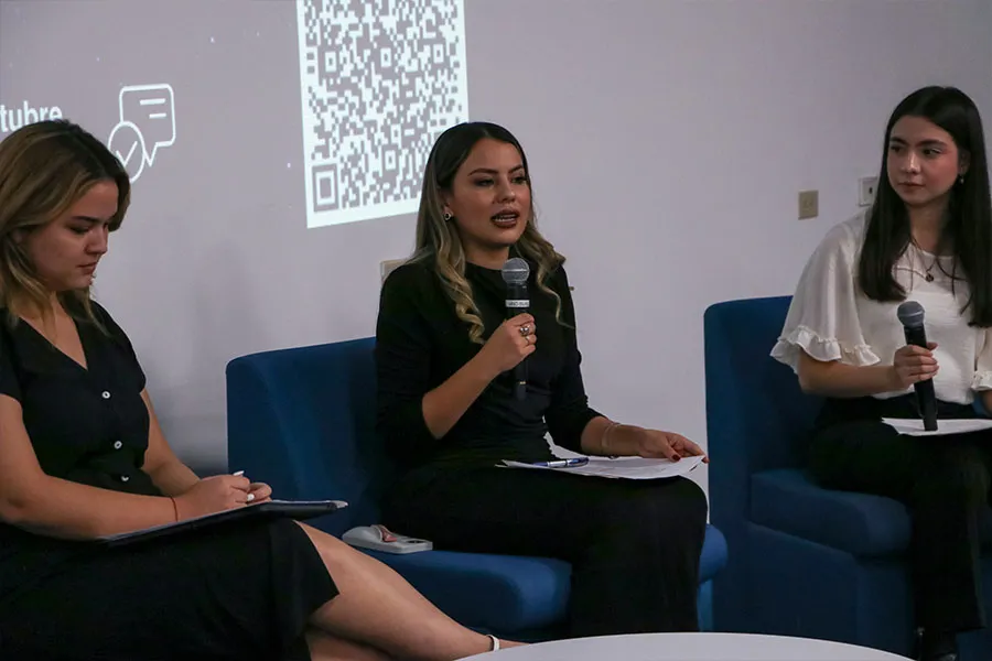 Tres mujeres sentadas en sillones azules mientras la persona del centro habla sosteniendo un micr&oacute;fono.