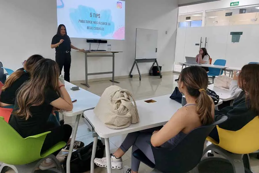 Mujer al frente de un sal&oacute;n de clases, habl&aacute;ndole a un grupo de alumnas. 