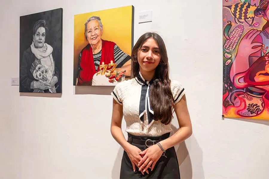 Mujer adolescente posando frente a una pintura que muestra a una mujer mayor con un pastel que tiene unas velas en forma del n&uacute;mero 77.
