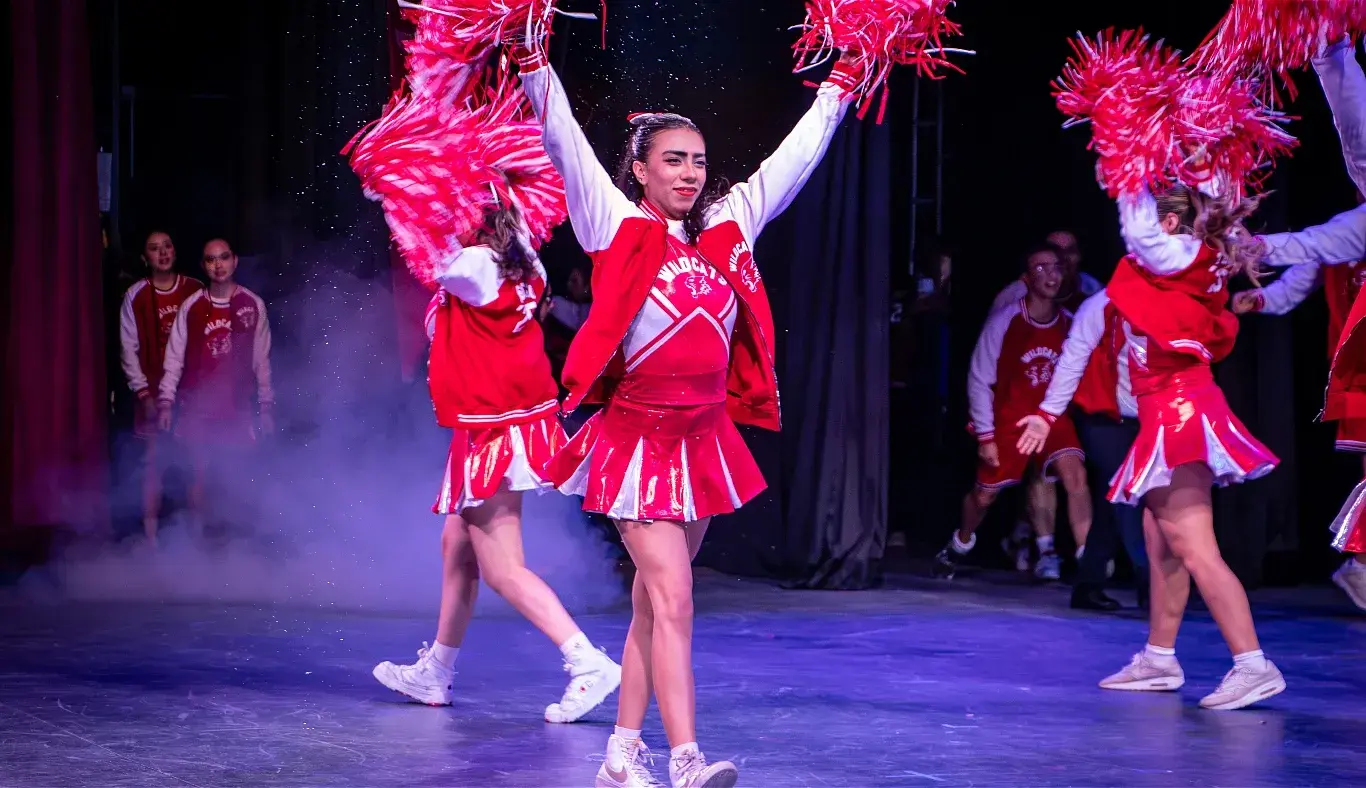 Estudiante caracterizada como porrista del equipo Wildcats realiza una coreografía con pompones durante el musical High School Musical: On Stage, en el escenario del campus Hidalgo.