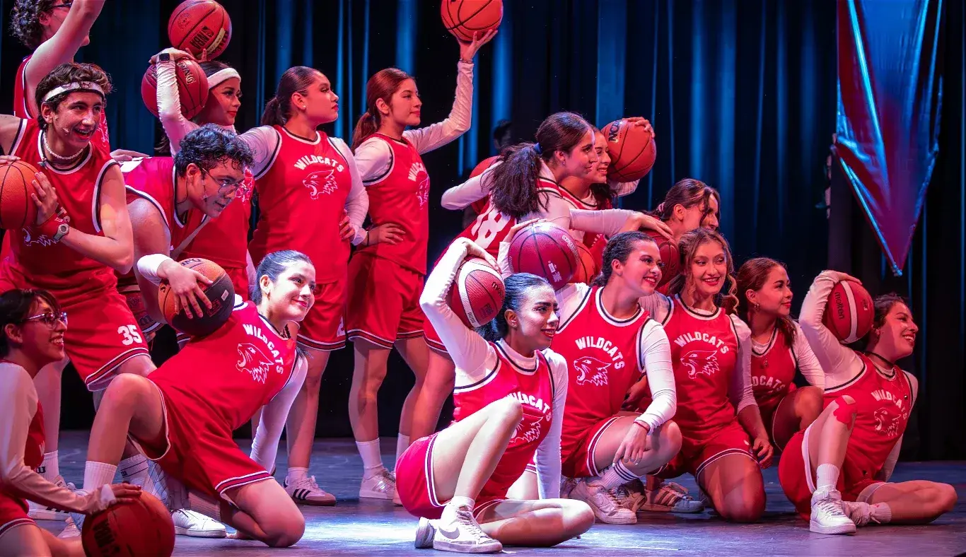 Estudiantes interpretan una coreografía con balones de baloncesto durante el número Get’cha Head in the Game del musical High School Musical: On Stage, con vestuario deportivo rojo.