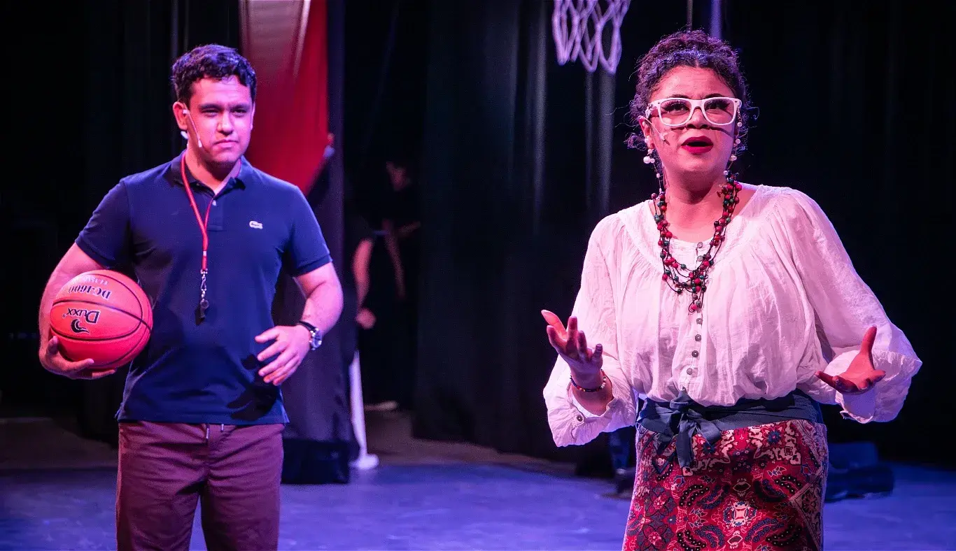 Estudiantes interpretan una escena de confrontación entre el entrenador Bolton y la señora Darbus durante el musical High School Musical: On Stage, con elementos representativos del deporte y el teatro.