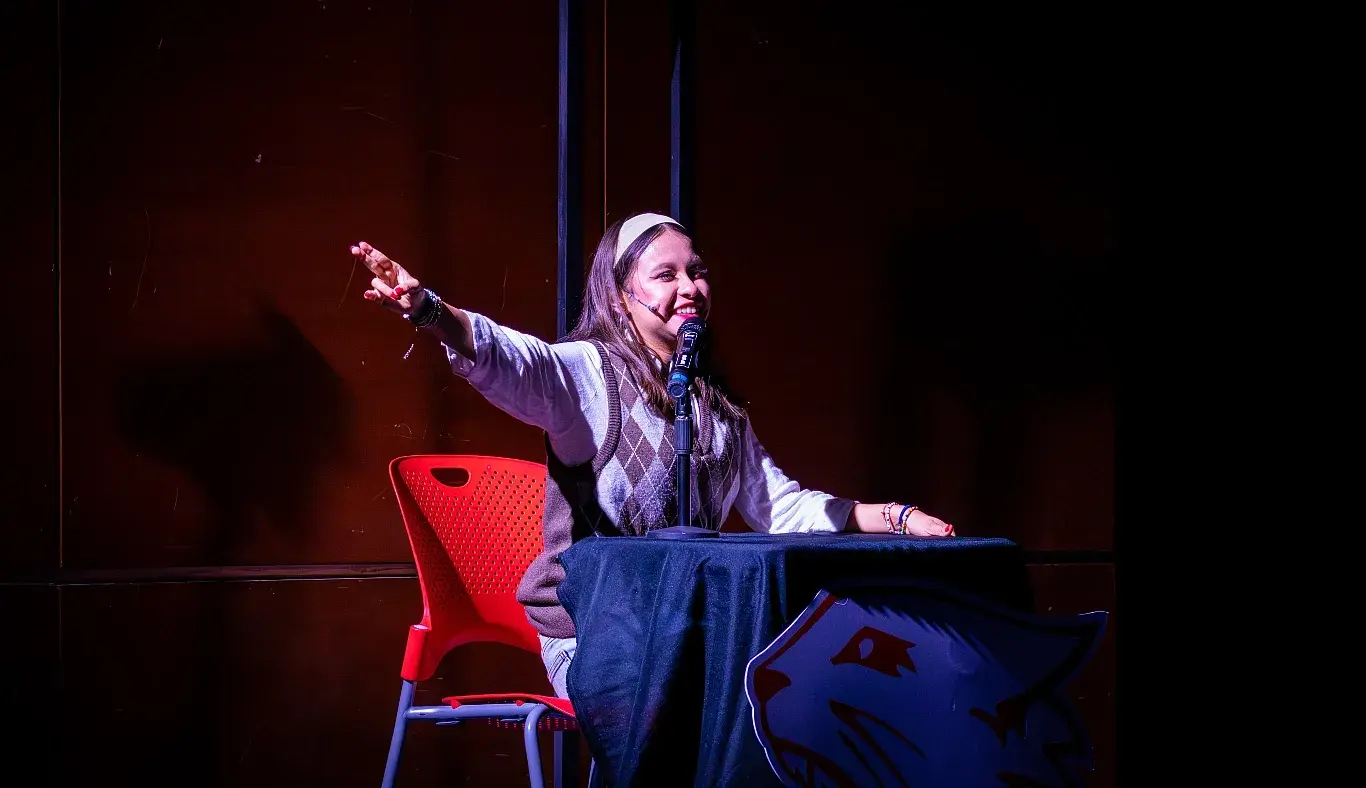 Estudiante caracterizada como Jackie Scott actúa como narradora escolar durante el musical High School Musical: On Stage, sentada frente a un micrófono con expresión entusiasta.