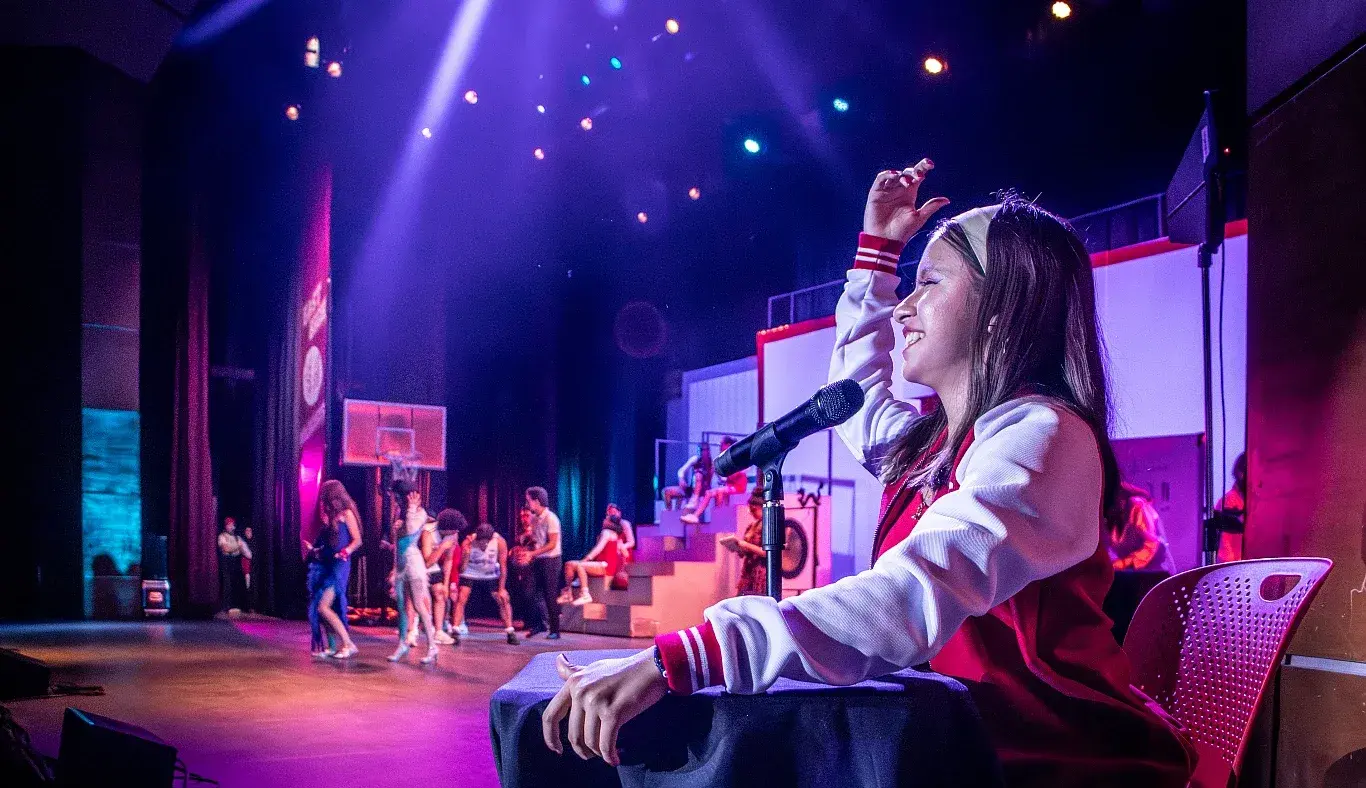 Estudiante caracterizada como Jackie Scott, la locutora escolar, conduce una escena desde la cabina con micrófono en High School Musical: On Stage, mientras en el fondo se desarrolla una coreografía.