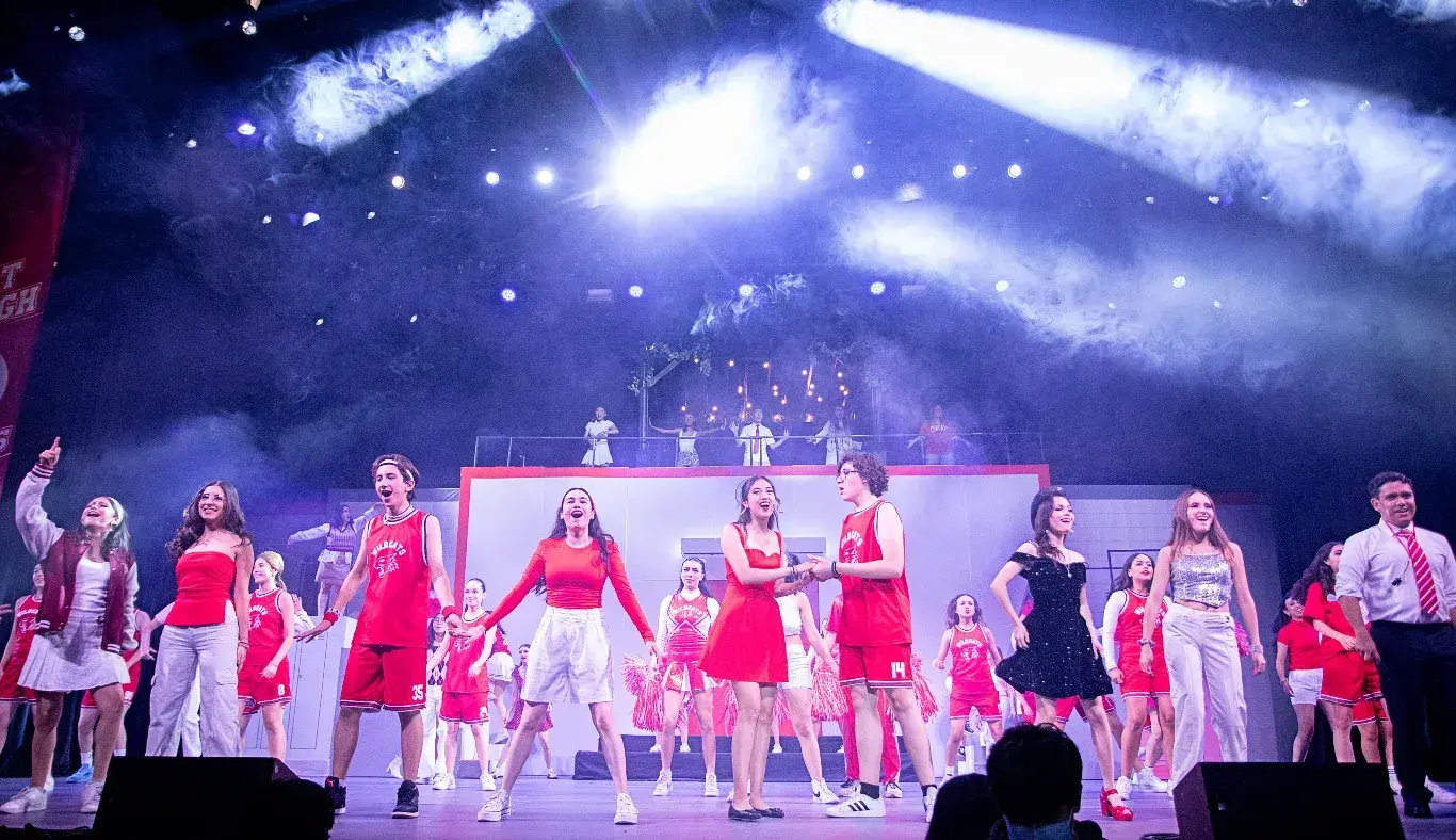 Elenco completo de High School Musical: On Stage interpreta el número final We’re All in This Together con escenografía iluminada, efectos de humo y participación grupal en el escenario principal.