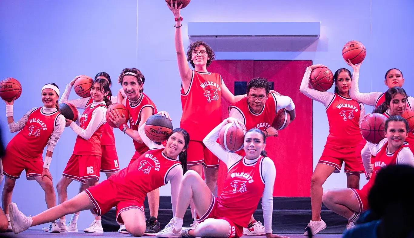 Estudiantes caracterizados como el equipo de baloncesto Wildcats posan con balones en escena durante High School Musical: On Stage, en una coreografía grupal del musical estudiantil.