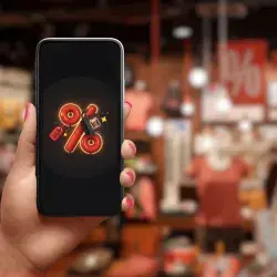 Tienda departamental al fondo de la imagen. En primer plano se observa una mano sosteniendo un celular, el cual refleja un signo de descuento