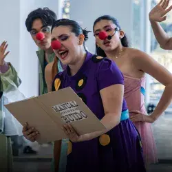 Actriz con nariz de payaso sostiene un folder y lee de él, mientras otros la ven.