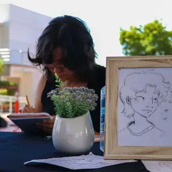 Chica dibujando sobre una mesa y enfrente de ella se ve un dibujo de una cara en un marco de madera. 