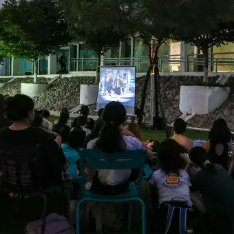 Grupo de estudiantes sentados en un jardín viendo una película proyectada sobre una pantalla blanca. 
