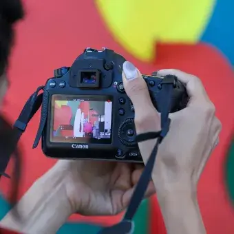 Vista sobre el hombro de unas manos sosteniendo una cámara y en su pantalla se ve la imagen de una escultura colorida. 