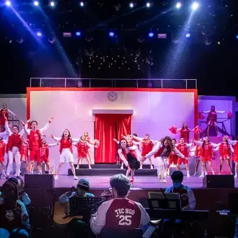El elenco completo interpreta el número final del musical High School Musical: On Stage, mientras al frente se distingue al director del ensamble musical guiando a los músicos del campus Hidalgo.