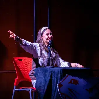 Estudiante caracterizada como Jackie Scott actúa como narradora escolar durante el musical High School Musical: On Stage, sentada frente a un micrófono con expresión entusiasta.