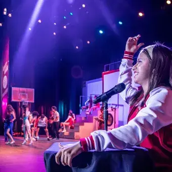 Estudiante caracterizada como Jackie Scott, la locutora escolar, conduce una escena desde la cabina con micrófono en High School Musical: On Stage, mientras en el fondo se desarrolla una coreografía.