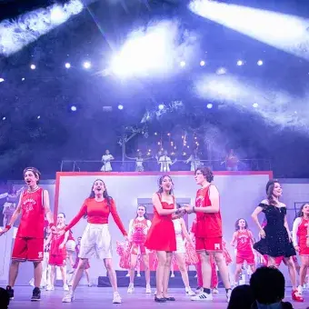 Elenco completo de High School Musical: On Stage interpreta el número final We’re All in This Together con escenografía iluminada, efectos de humo y participación grupal en el escenario principal.