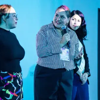 Tres mujeres paradas frente a un salón, mientras la de en medio sostiene un micrófono.