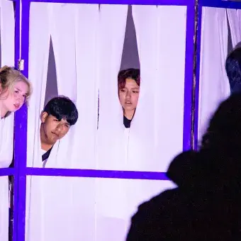 5 jóvenes se asoman entre cortinas por una ventana. 