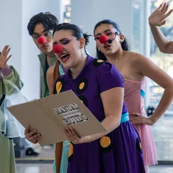 Actriz con nariz de payaso sostiene un folder y lee de él, mientras ot