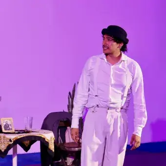Hombre vestido de blanco con sombrero negro en un escenario con luz rosa.
