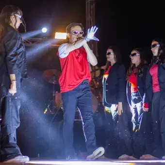 Cantante caminando hacia el frente del escenario rodeado de bailarinas a cada lado.