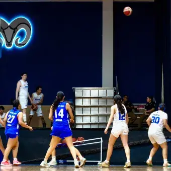 Estudiante de básquetbol femenil ensaya un tiro de larga distancia con técnica de lanzamiento mientras todas las jugadoras observan.